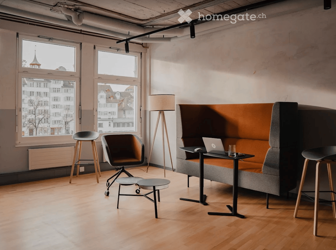 Modern, minimalist room with large windows, a grey sofa, a laptop on a small table, two stools, a chair, and a floor lamp.