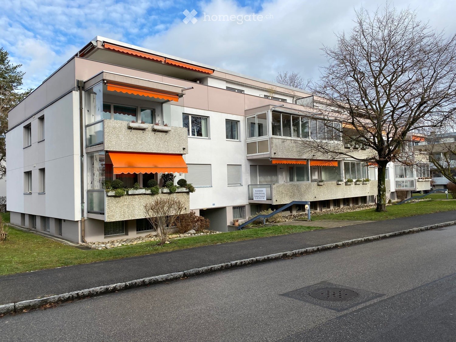 Modern two-story residential building with balconies, orange awnings, and small garden areas.