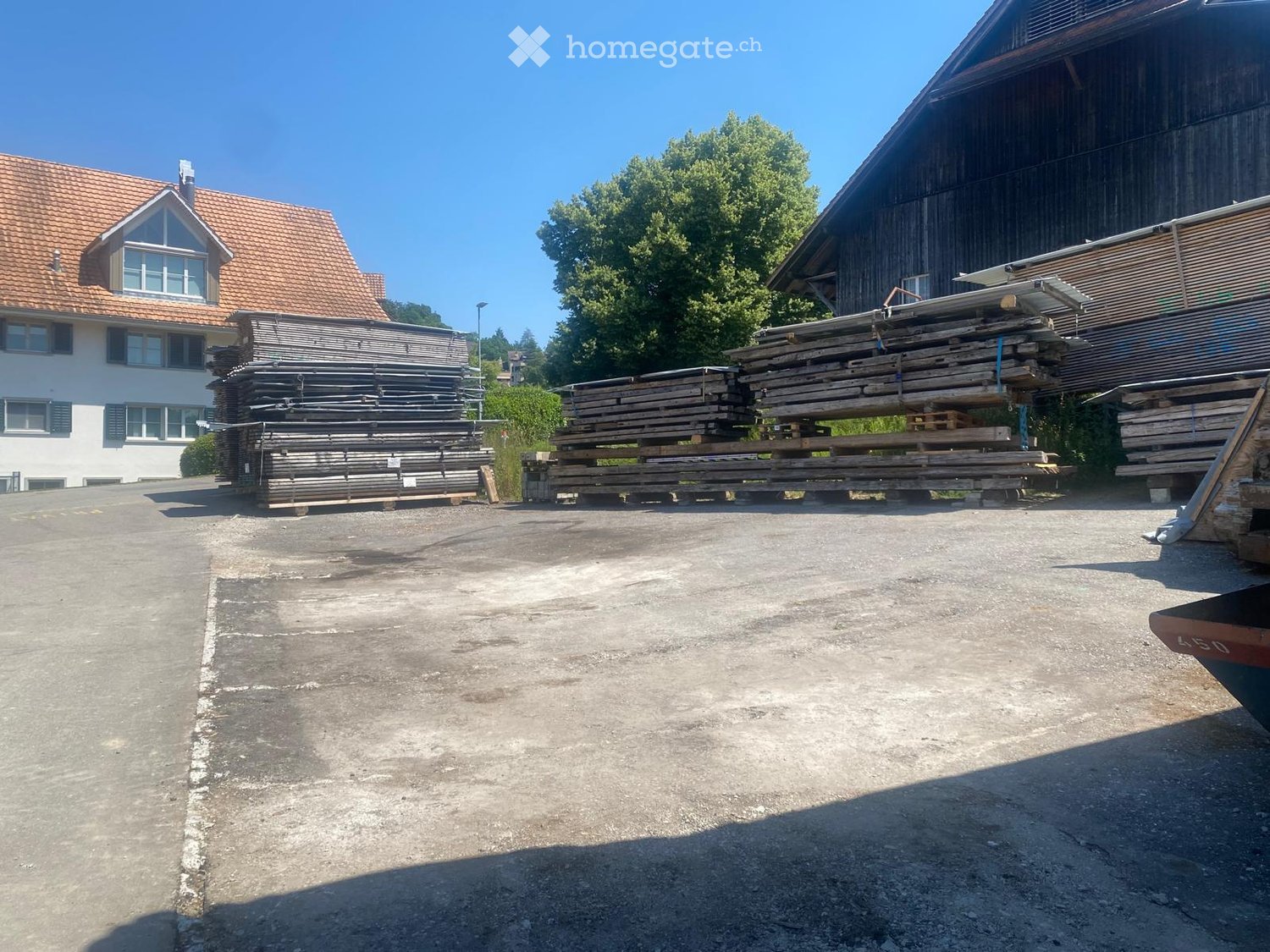 Outdoor area with a concrete floor, stacks of wooden planks, and a large wooden building in the background.
