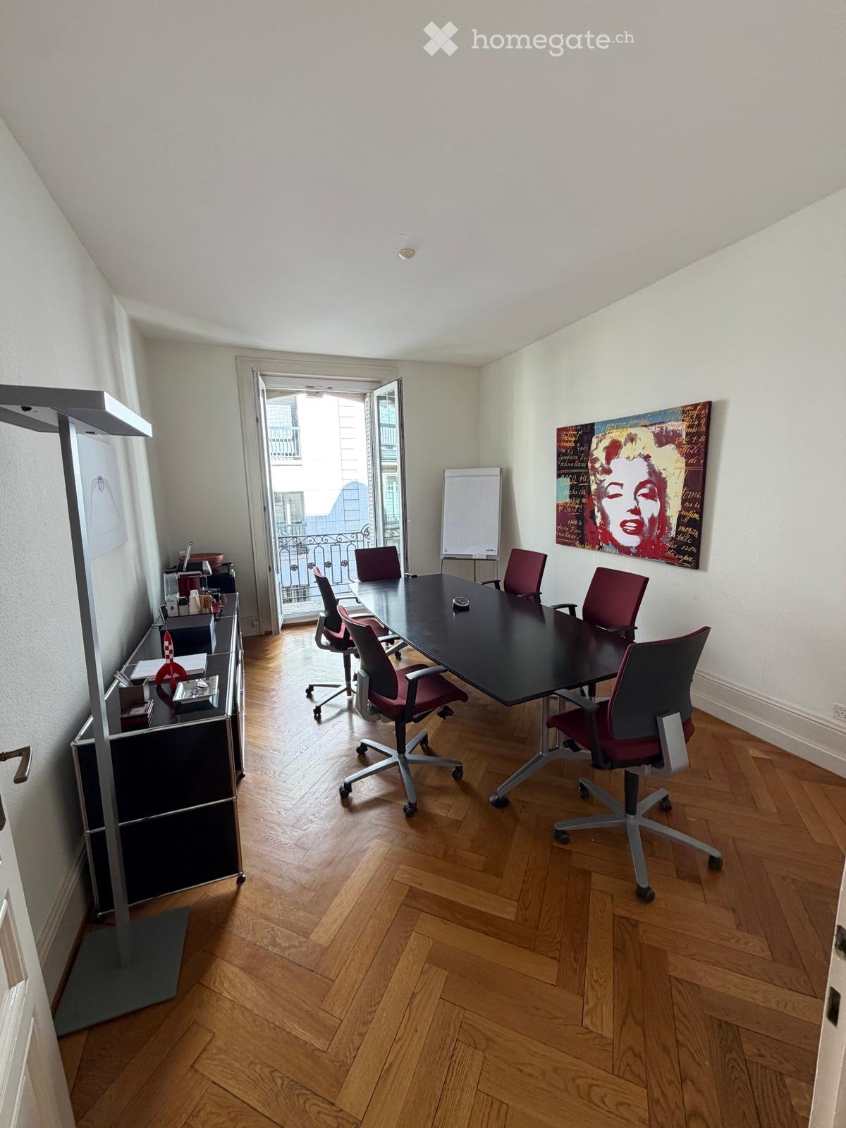 Office space with wooden flooring, a large black conference table surrounded by red chairs, a cabinet with office supplies, and a large screen on the wall. An open door leads to a balcony.