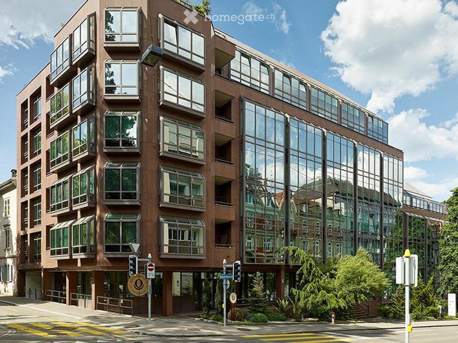Multi-story building with large windows and balconies, modern design, located on a street with traffic signs and greenery.