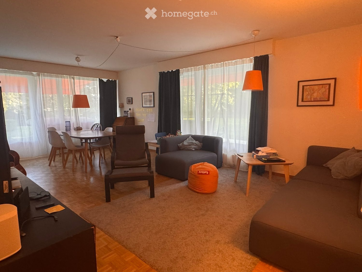 A modern living room with a sofa, a chair, a small table, a round stool, a dining table with chairs, and a TV stand with a screen. The room has large windows with curtains and is well-lit with hanging lamps.