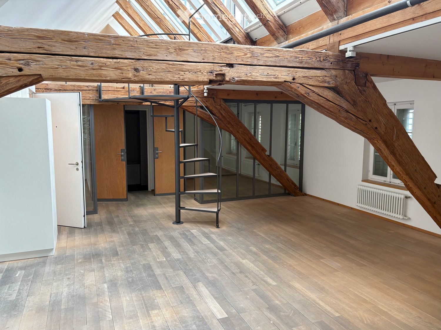 Unfurnished Room. Open, high-ceilinged room with exposed wooden beams, wooden flooring, a modern staircase, and large windows allowing natural light.