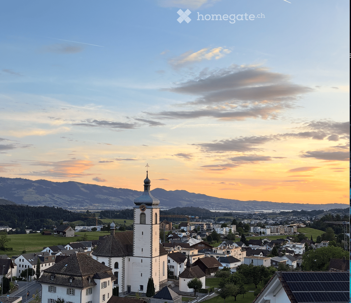 A picturesque town view at sunset, featuring a prominent white church tower with a blue dome and clock, surrounded by houses and green fields, with mountains in the background.