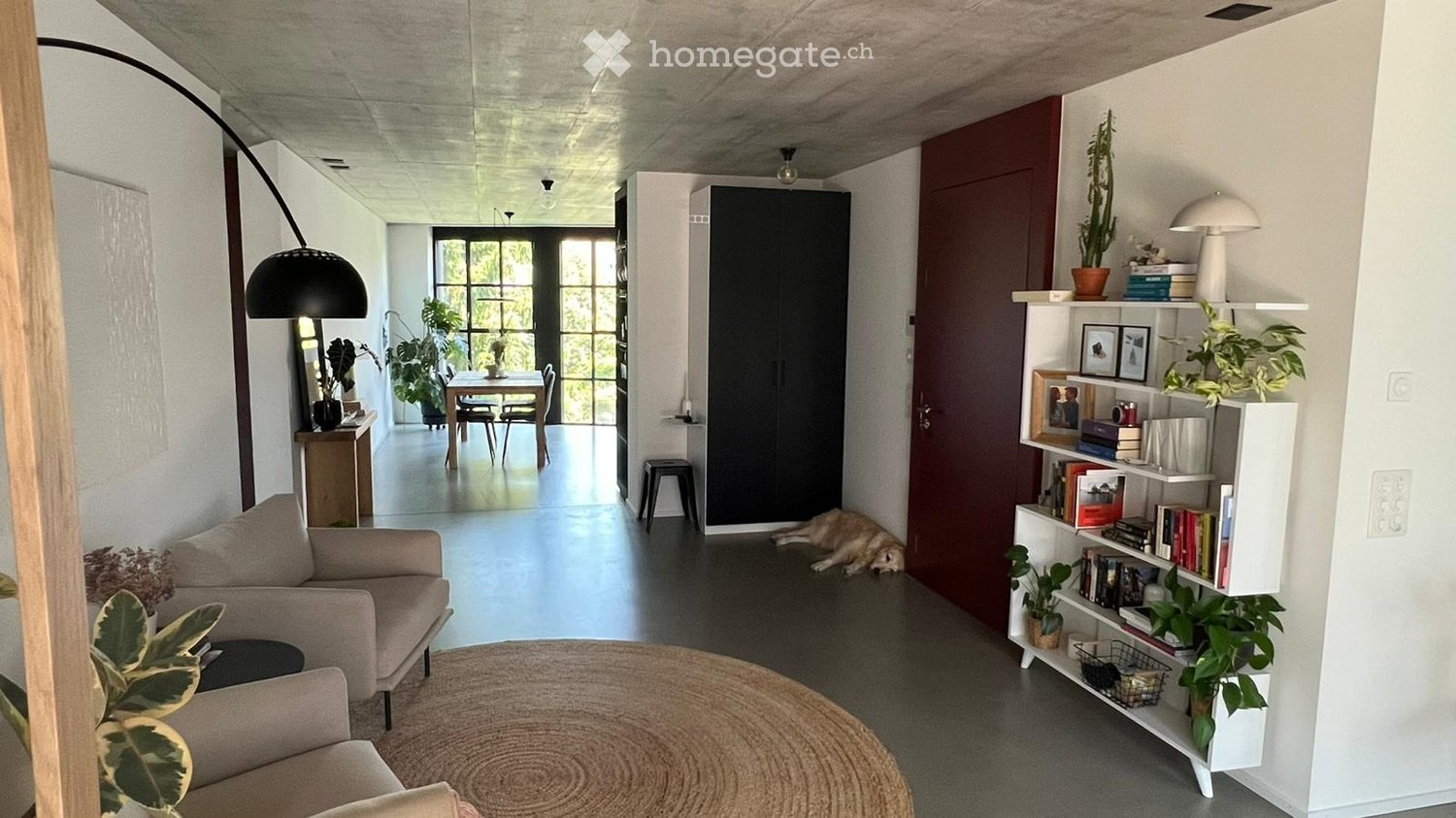 Modern living room with a sofa, coffee table, bookshelf, floor lamp, dining table, chairs, and a small stool. The space is well-lit with large windows and features several potted plants.