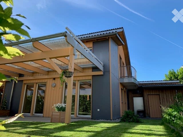 Modern two-story house with a wooden pergola, large windows, and a balcony on the upper level.