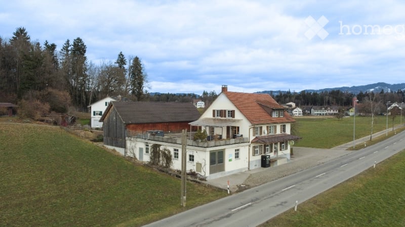 A two-story house with a red-tiled roof, white walls, and a balcony terrace. The house is surrounded by a grassy area and trees, with a road running alongside.