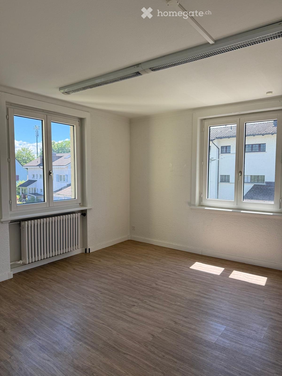 Empty room with wooden flooring, white walls, large windows, radiator, and visible electrical outlets.