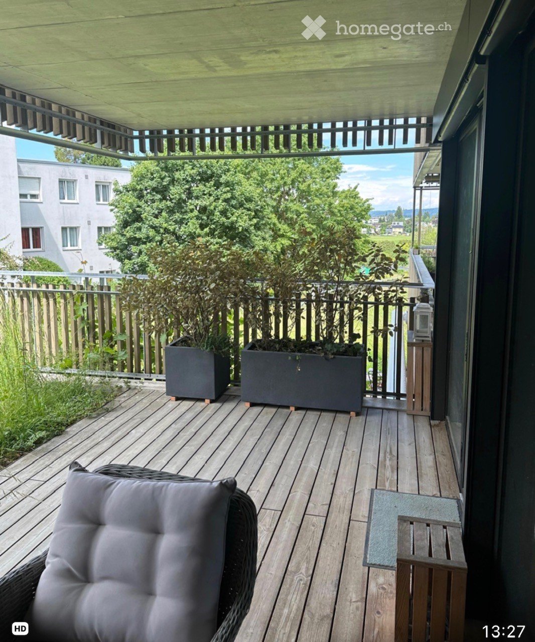 Wooden decked balcony with a wicker chair, small wooden table, and two large planters. The balcony has a wooden railing and a covered roof, with a scenic view of greenery and buildings in the background.