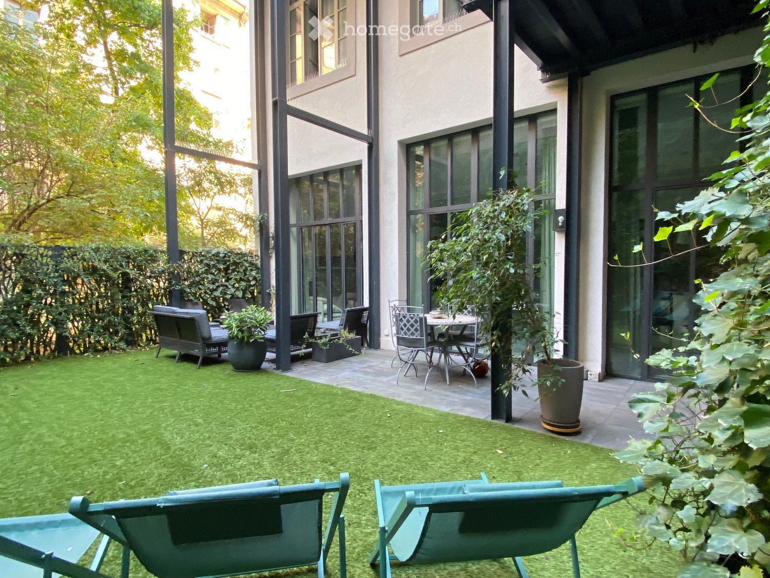 Garden area with artificial grass, lounge chairs, sofa, dining table, and potted plants near a modern building with large windows.
