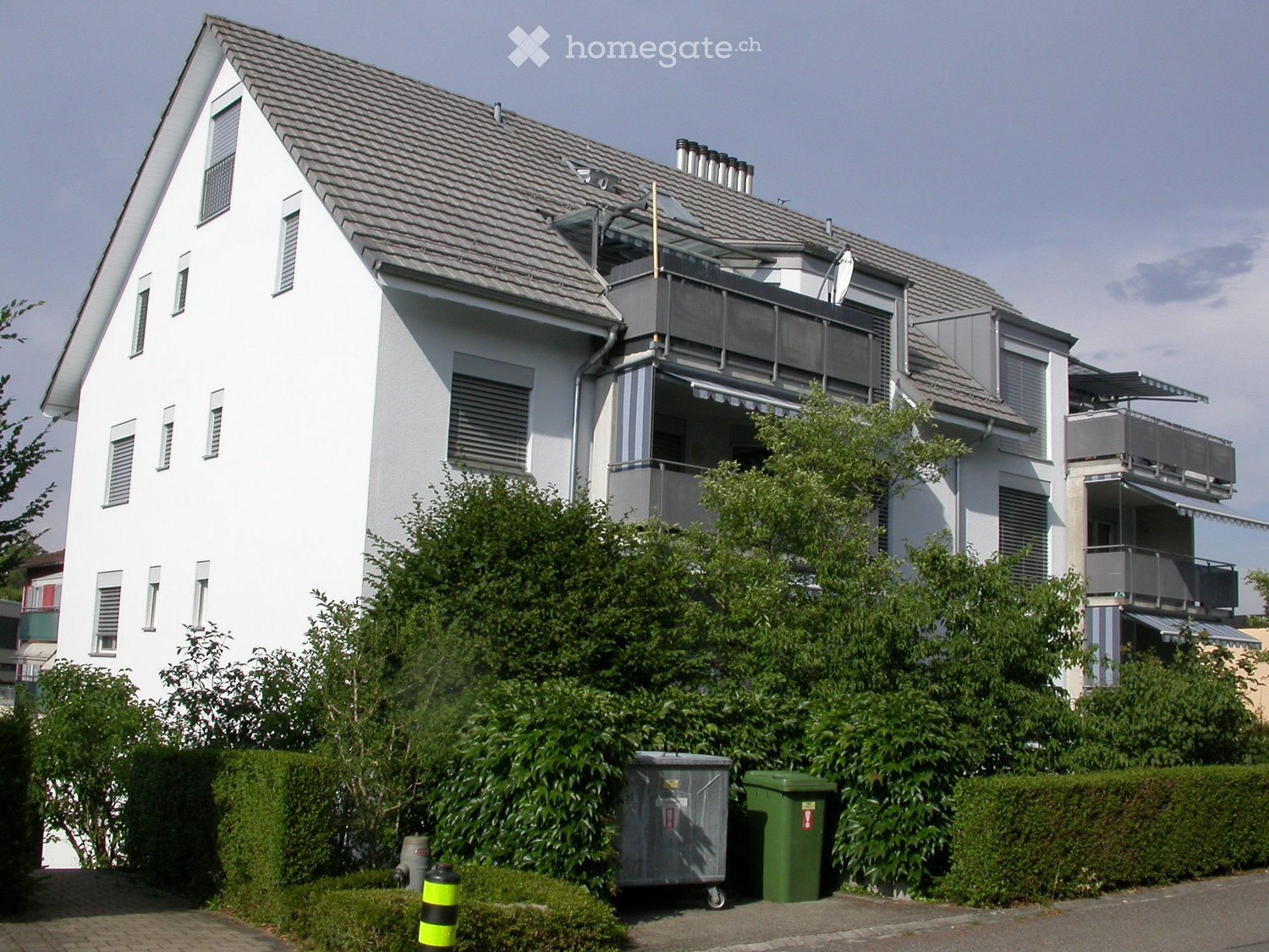 Modern two-story house with white exterior, multiple windows, balconies, and surrounding greenery.