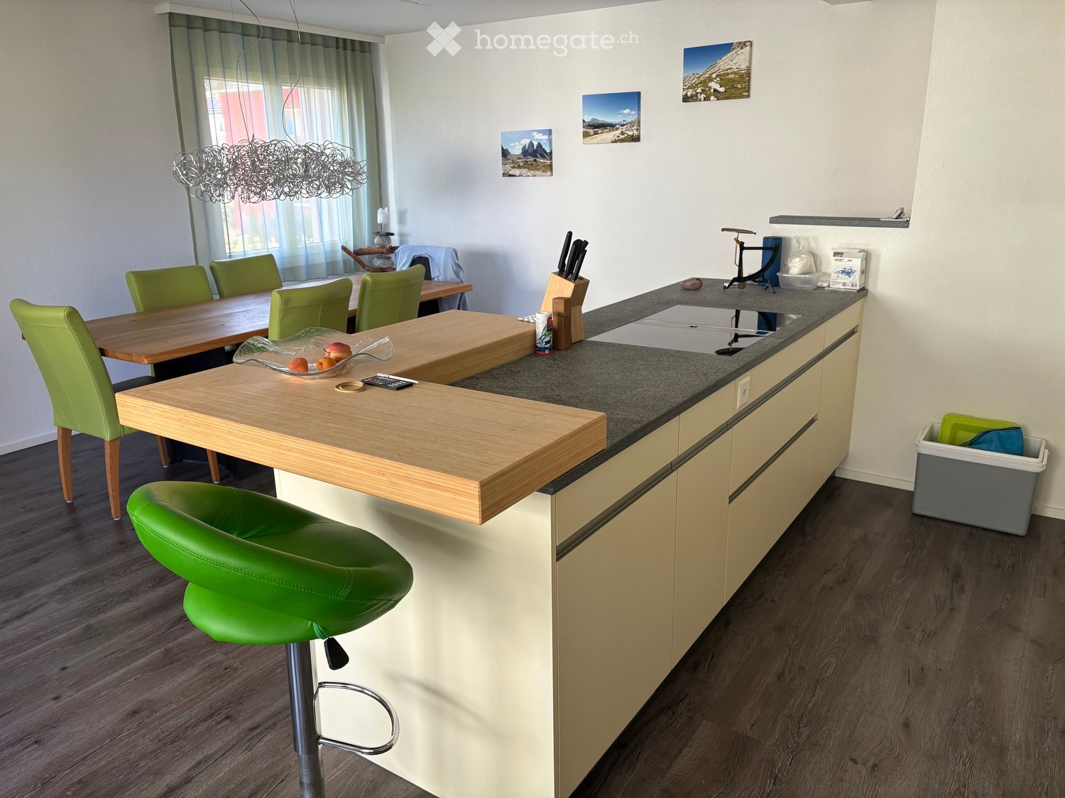 Modern kitchen with light wood cabinets, dark granite countertop, and a wooden dining table with green chairs. Features a bar stool, knife block, and various kitchen items.