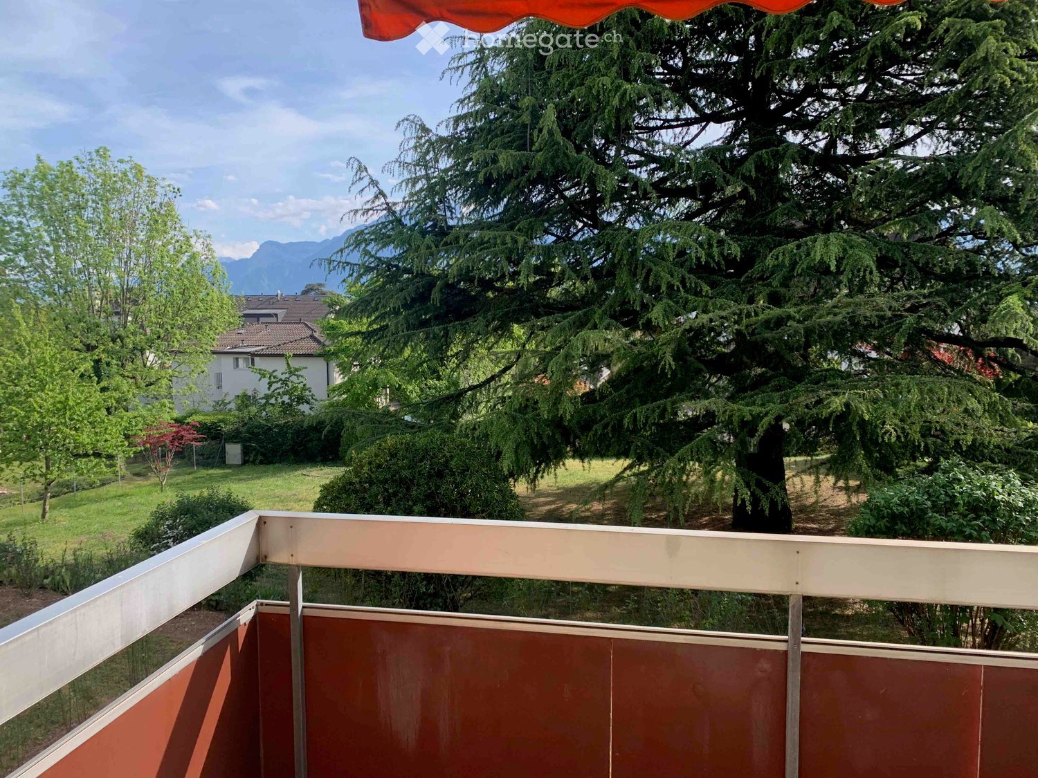 Balcony with wooden railing, overlooking a green garden and trees, with a distant view of mountains.