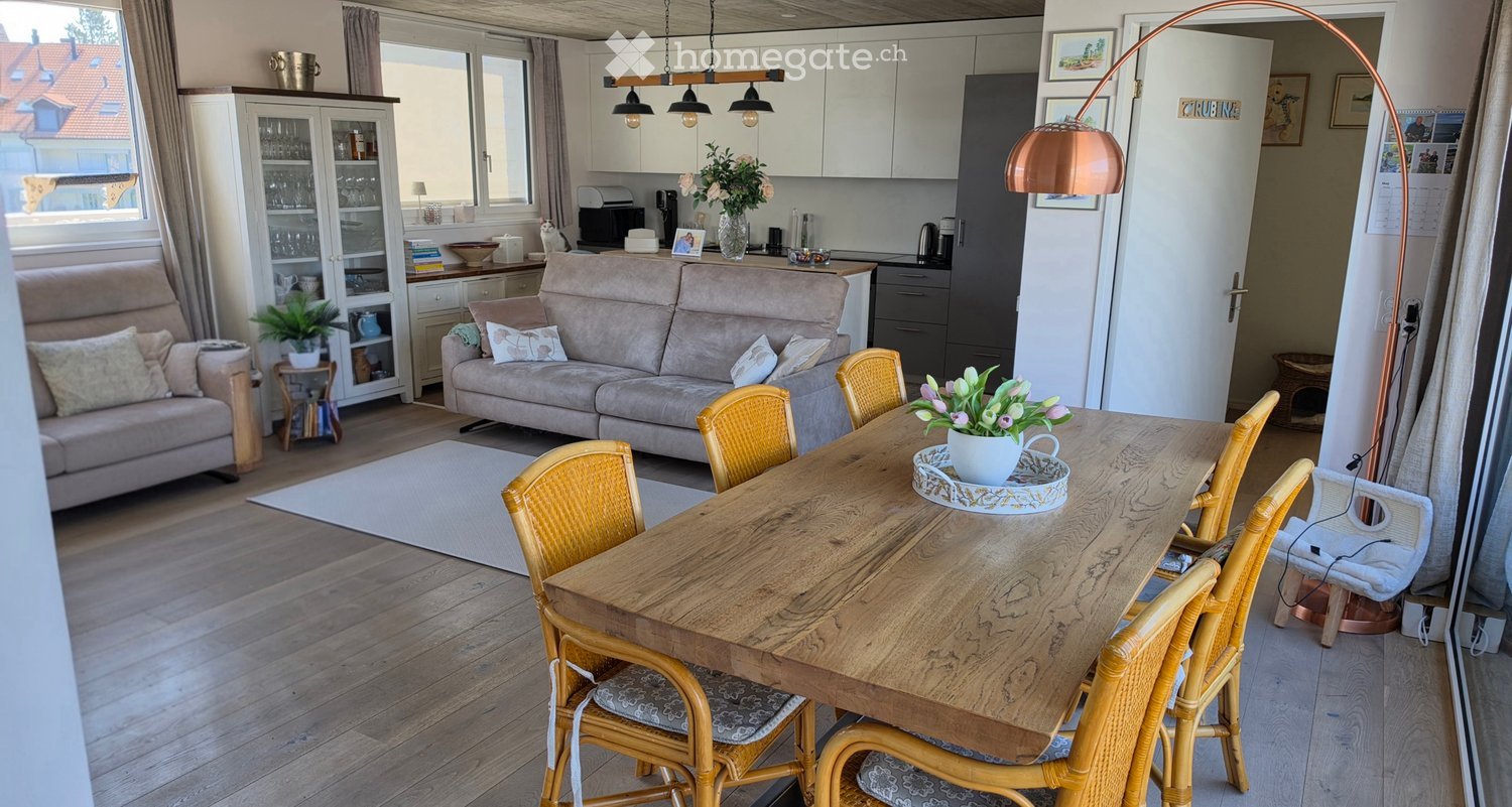 Bright living and dining area with wooden flooring, a large sofa, a dining table with chairs, a floor lamp, and a cabinet with glass doors.