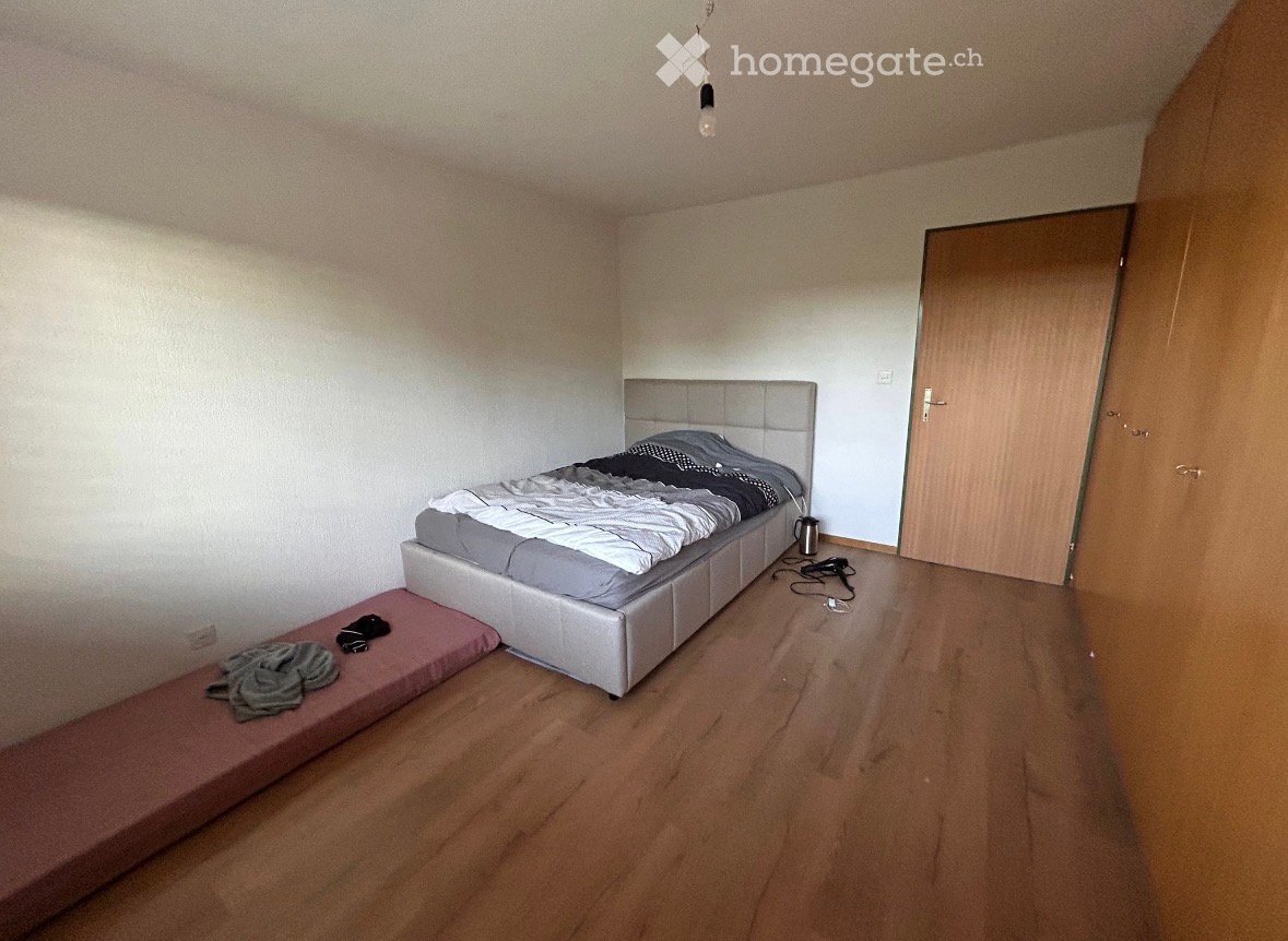 Bedroom with a grey upholstered bed, a matching nightstand, and a small bench at the foot of the bed. Wooden flooring and white walls.