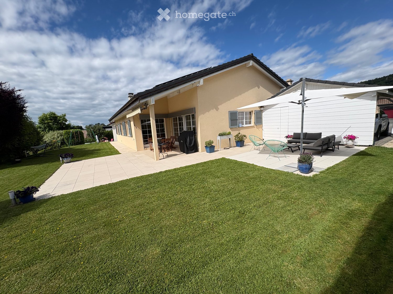 A well-maintained garden with a lawn, patio seating area, outdoor furniture, and a covered patio connected to a house.