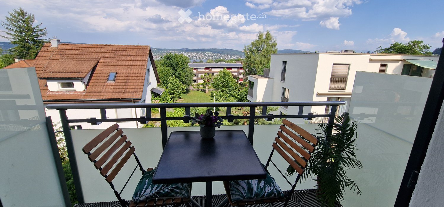 Balcony with a small table, two chairs, and a flower pot, overlooking a residential area with houses and greenery.
