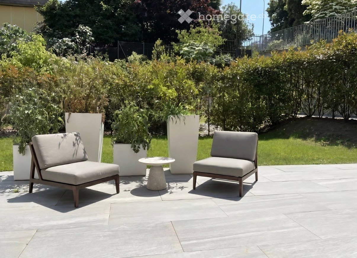 A modern outdoor seating area with two beige chairs and a small round table on a concrete patio, surrounded by lush greenery and tall white planters.