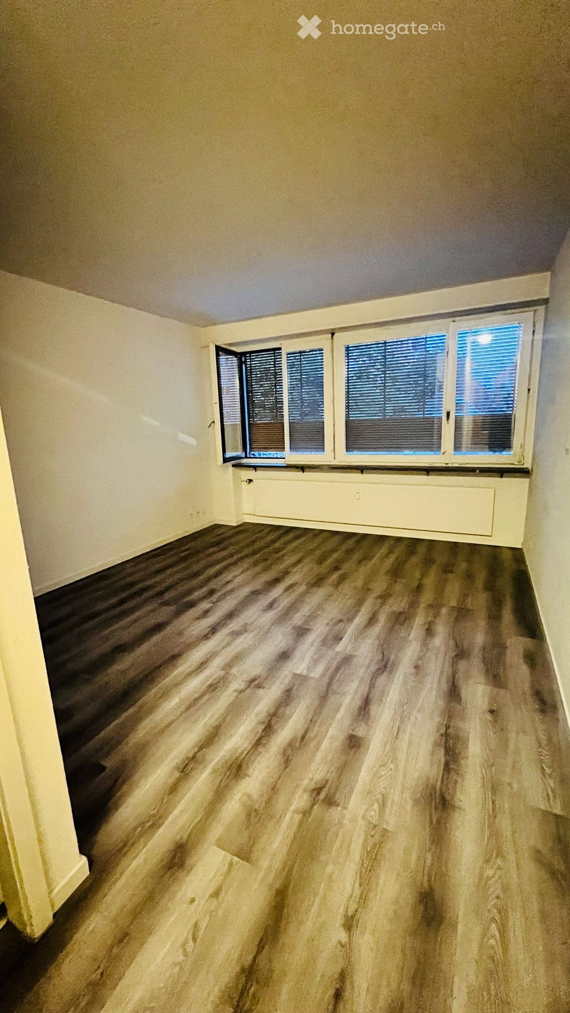 Empty room with wooden flooring, white walls, and large windows with blinds. No furniture or equipment present.