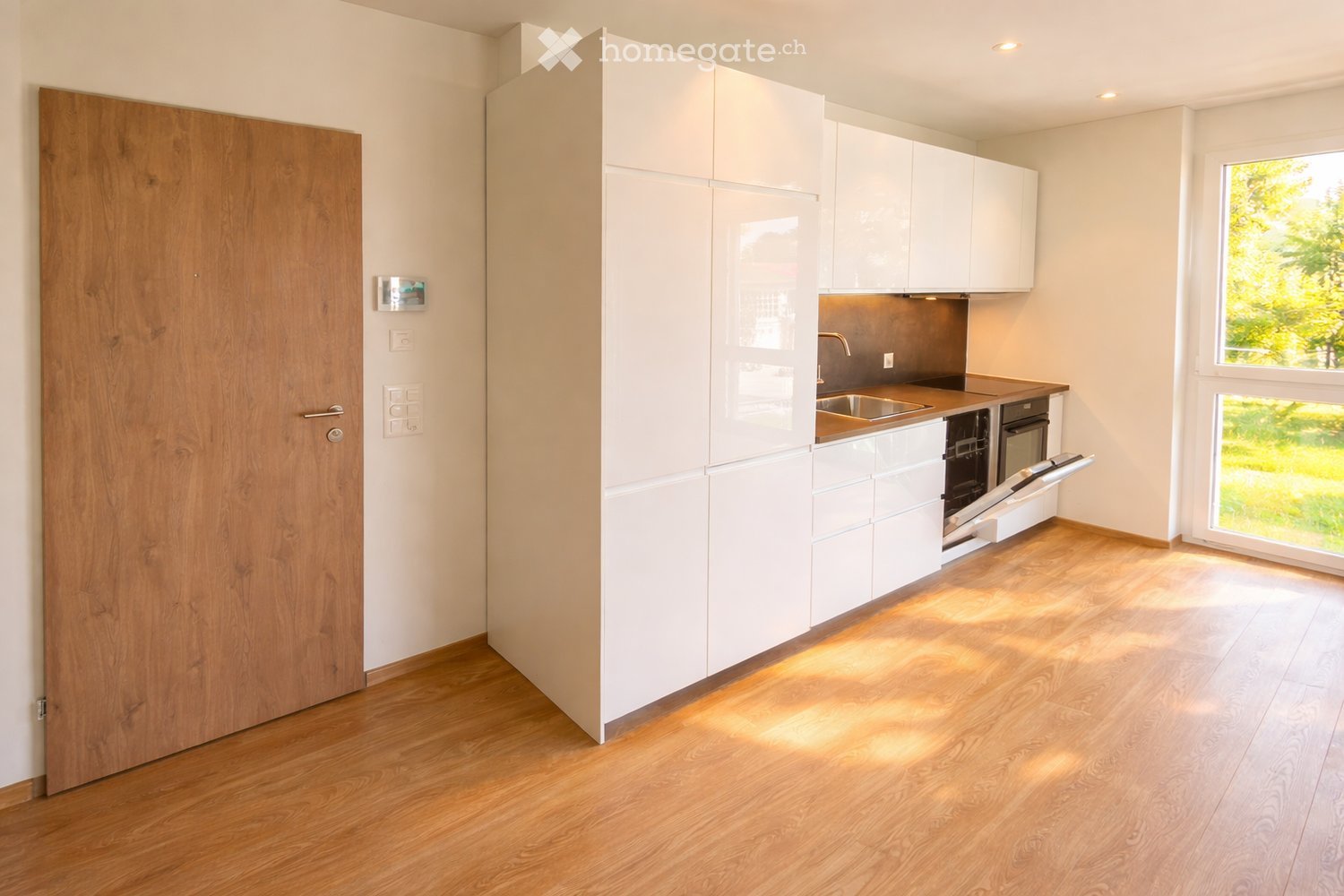 Modern kitchen with white cabinets, wooden floor, stainless steel sink, oven, and dishwasher. Large windows provide natural light.
