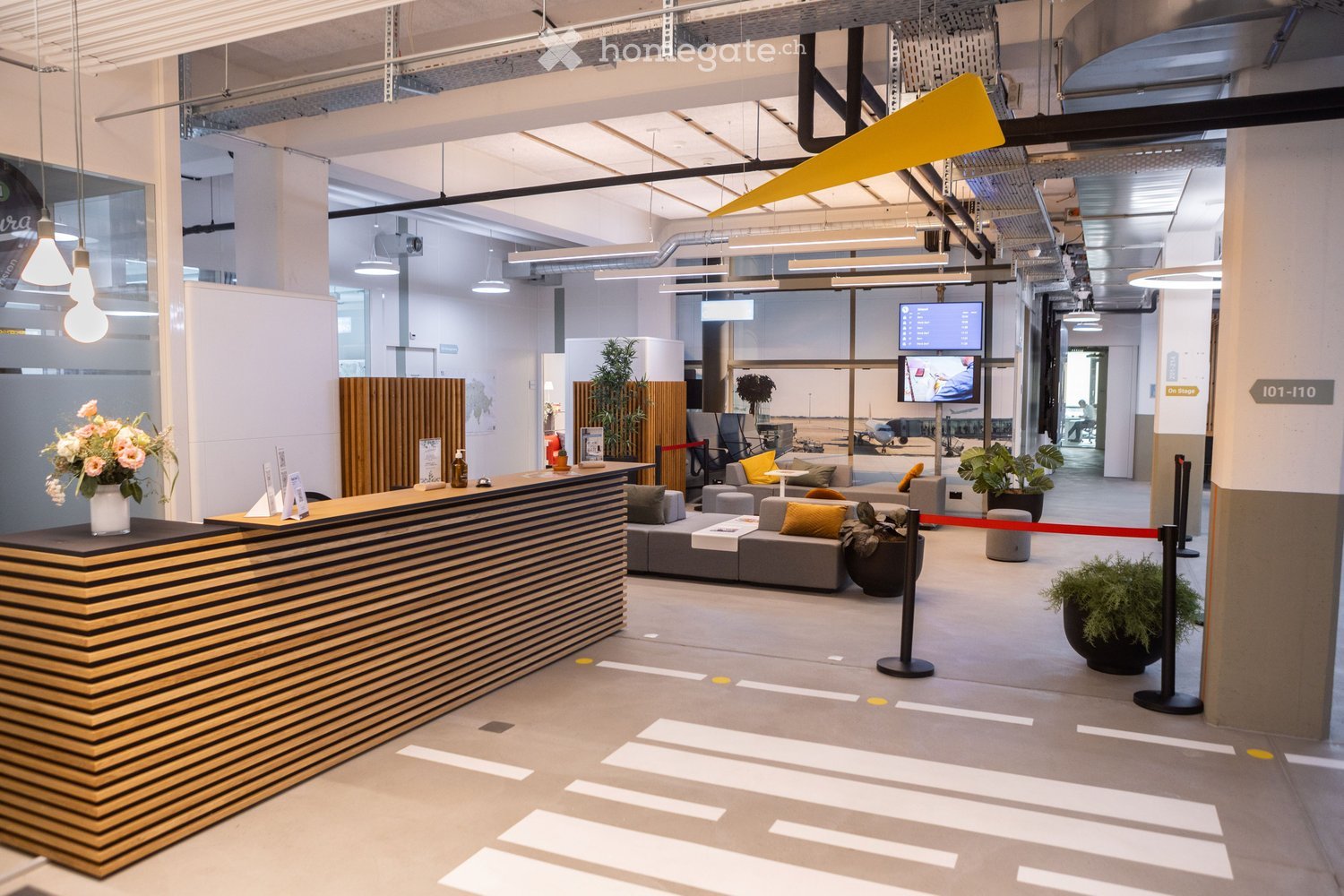 Modern reception area with a curved wooden reception desk, seating area with sofas and chairs, potted plants, and a television screen displaying information.