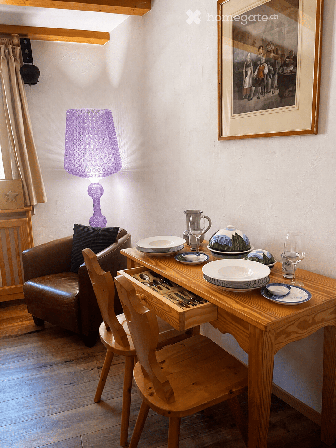 A cozy, well-furnished room with a wooden dining table set for a meal, a brown leather sofa, a wooden chair, and a decorative purple lamp. The room features a framed picture on the wall and a curtained window.