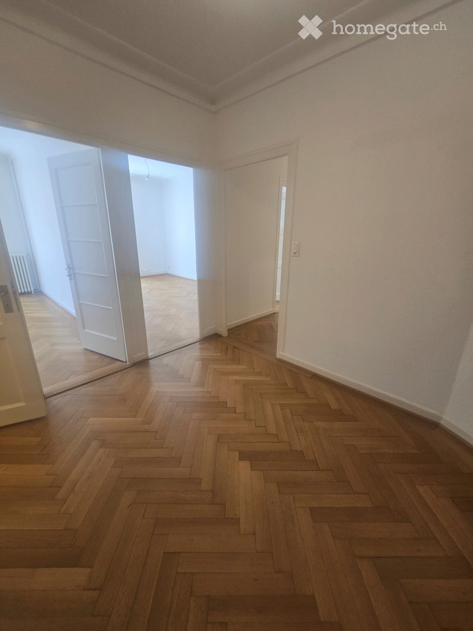 Empty room with wooden herringbone flooring, white walls, and an open door leading to another room.