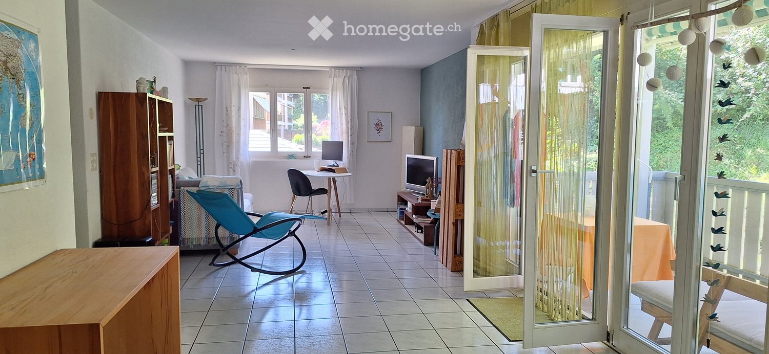 A well-lit living room with tiled flooring, large windows, and a sliding glass door leading to a balcony. The room features a blue rocking chair, a wooden cabinet, a small table with a screen, and a second screen on a stand. There is also a small table wi
