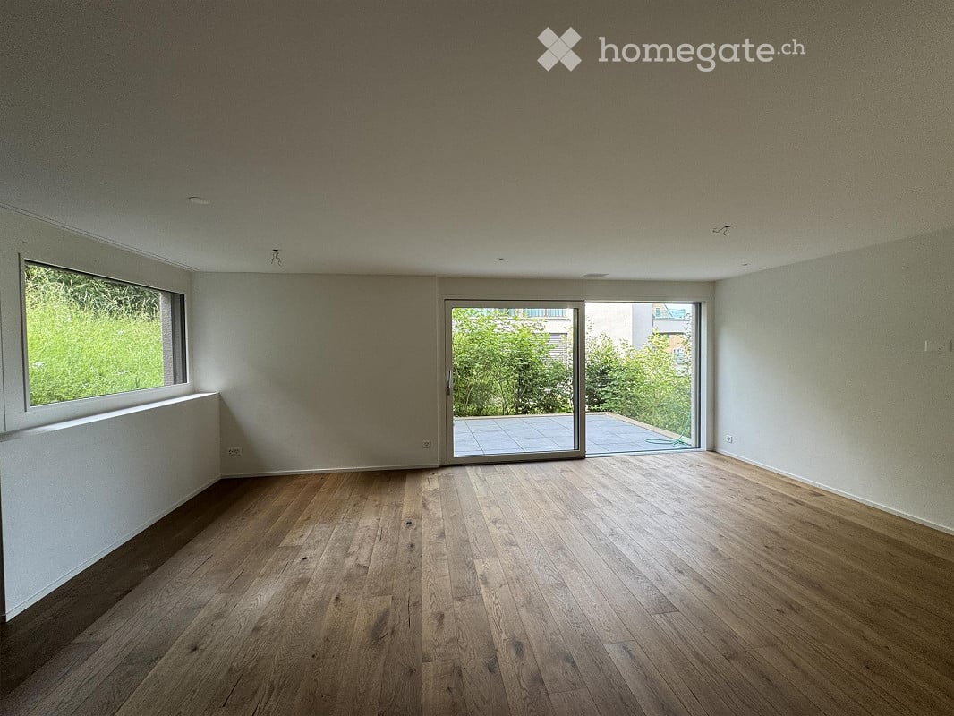 Empty room with wooden flooring, large windows, and sliding glass doors leading to a patio.