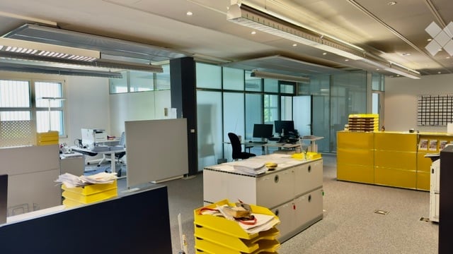 Modern office space with desks, chairs, computers, filing cabinets, and whiteboards. Bright lighting and large windows.