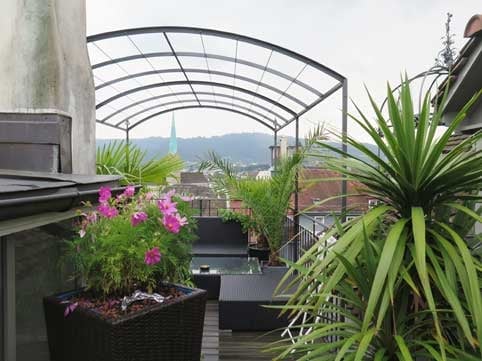 Rooftop terrace with a metal pergola, potted plants, and a view of the city and mountains.