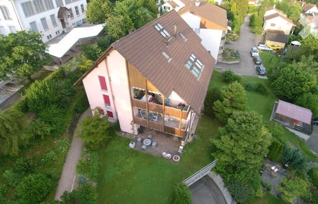 Aerial view of a modern two-story house with a brown tiled roof, multiple windows, and a spacious garden area with outdoor seating and a small patio.