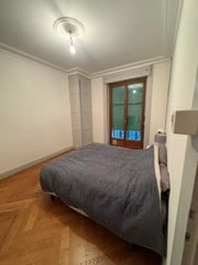 A modern bedroom with a wooden floor, white walls, and large windows with green-tinted glass. The room features a grey duvet on the bed, a bedside table with a lamp, and a ceiling light fixture.