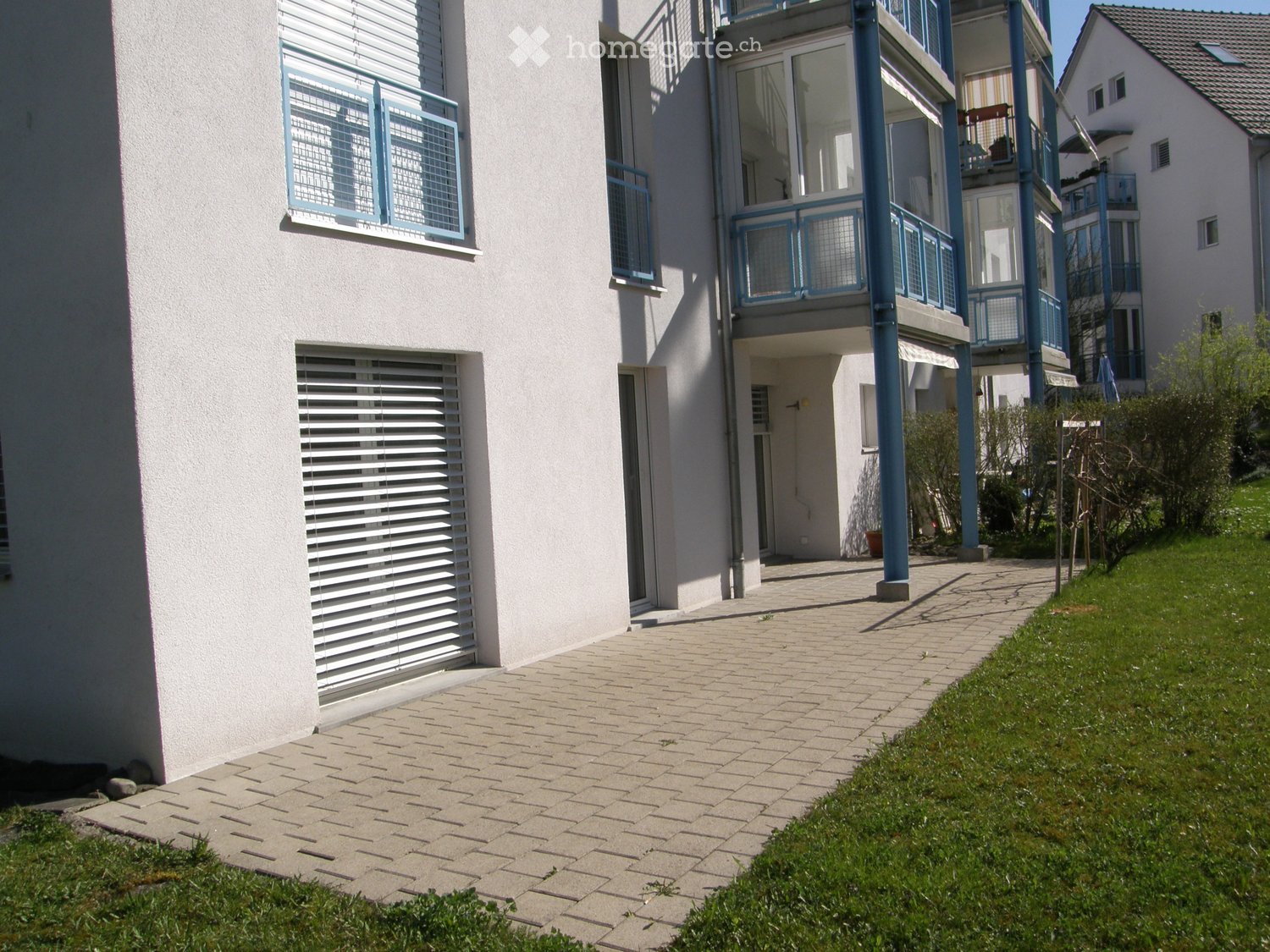 Modern multi-story residential building with white walls, blue-framed windows, and balconies. The entrance features a paved walkway leading to a door, with green grass and small plants on either side.