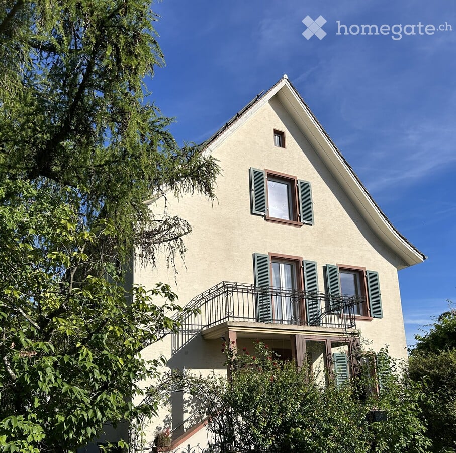 Two-story house with beige walls, green shutters, and a balcony on the first floor.