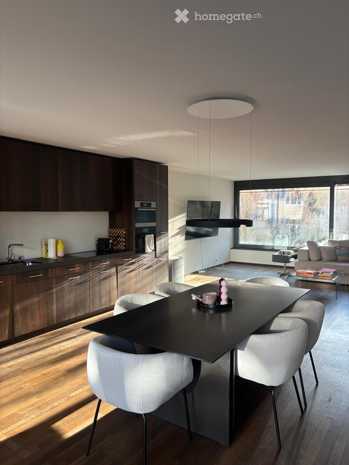 Modern open-plan living and dining area with a sleek black dining table, white chairs, a grey sofa, and a kitchen area with dark wooden cabinets and stainless steel appliances.