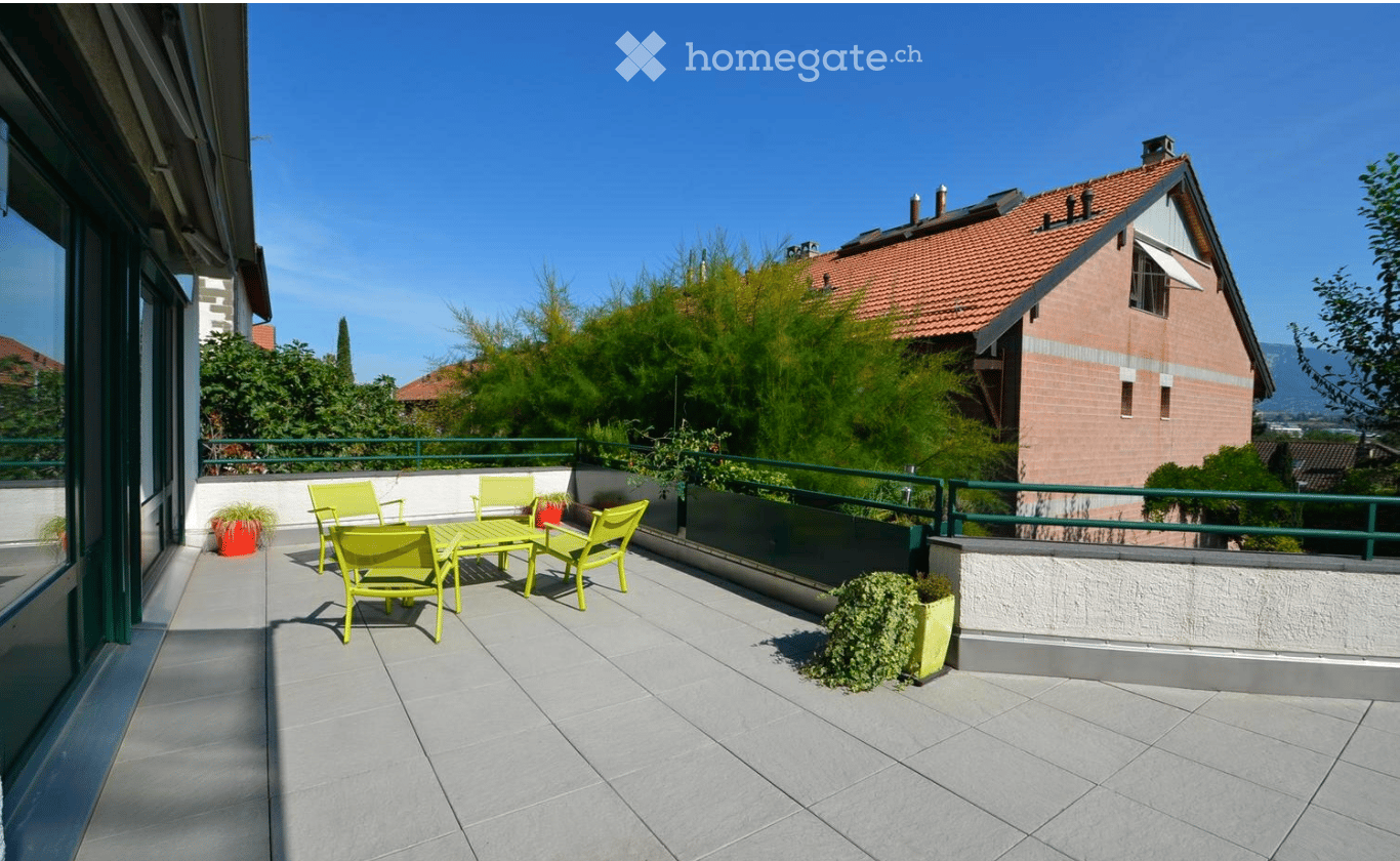 Balcony with tiled floor, green metal railing, yellow table and chairs, potted plants, and a view of a residential area with trees and a house.