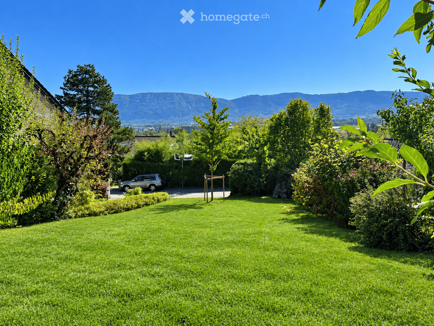 Well-maintained garden with a lush green lawn, surrounded by trees, shrubs, and a parked car in the background. Distant mountains are visible under a clear blue sky.