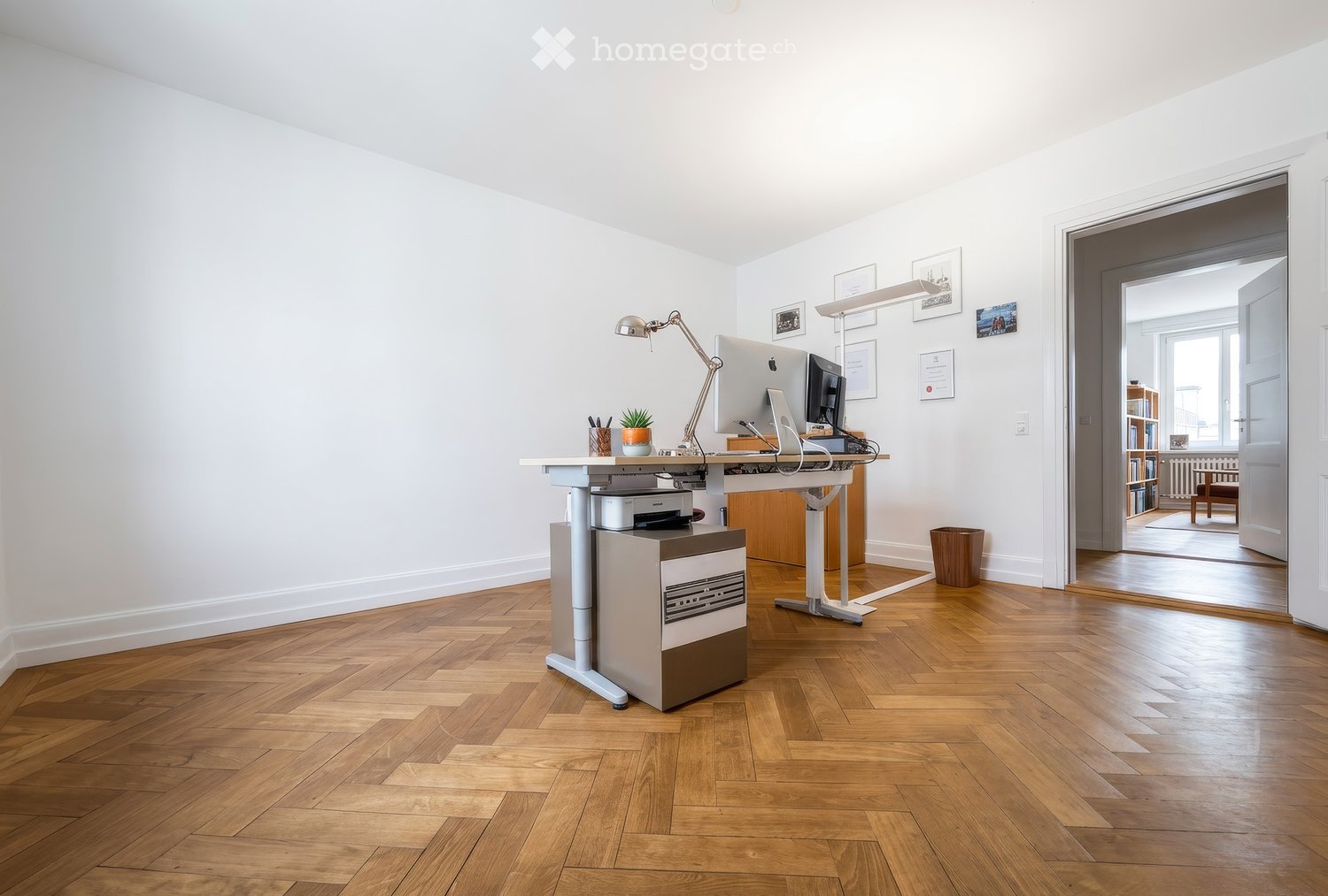 Modern office space with wooden flooring, white walls, and a standing desk setup. Includes a computer, printer, chair, lamp, small plant, and picture frames on the wall. An open doorway leads to another room with visible furniture.