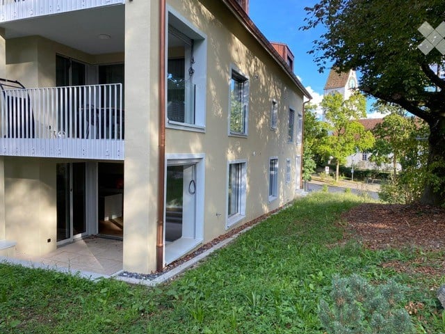Two-story residential building with a balcony on the upper floor, surrounded by greenery and a paved pathway.