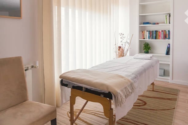 A well-lit room with a massage table, a chair, a bookshelf with books and a small plant, and a decorative rug on the floor.