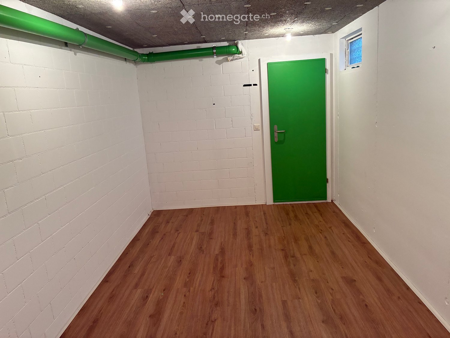 Empty hallway with white brick walls, wooden flooring, green doors, and a small window.