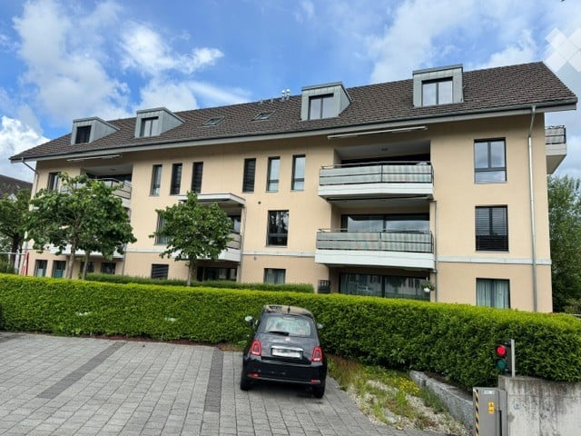 Modern multi-story residential building with balconies, large windows, and a neatly trimmed hedge in front. A small car is parked in the driveway.