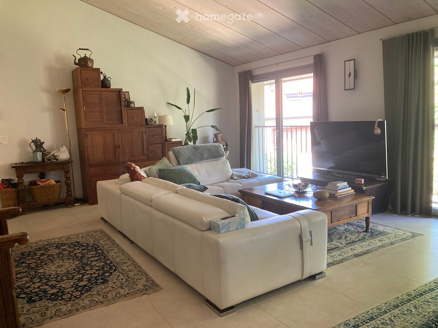 Bright living room with white leather sofa, coffee table, TV, wooden cabinet, and large windows opening to a balcony.