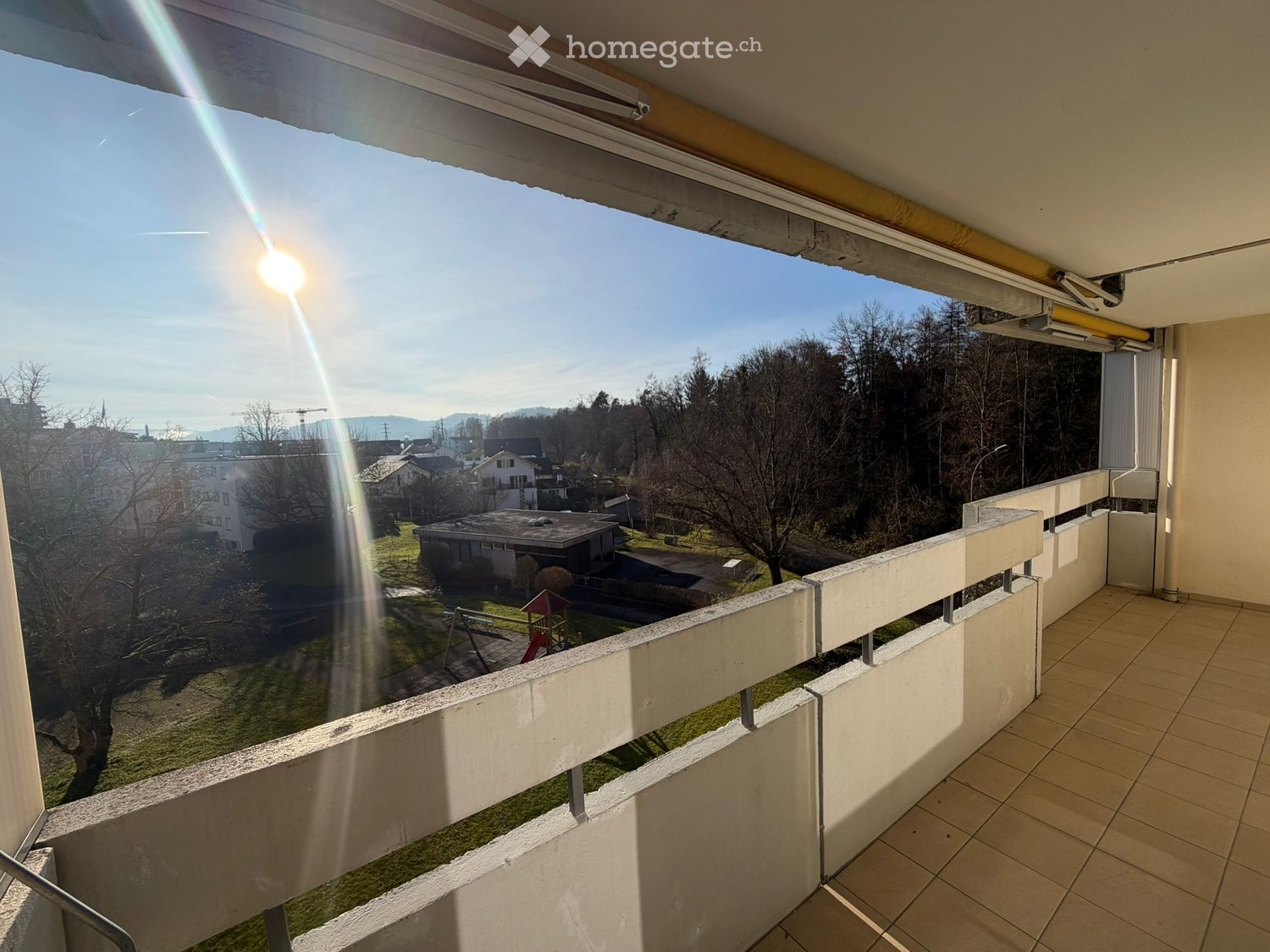 Balcony with tiled floor, white railing, overlooking a park with trees, houses, and a playground.