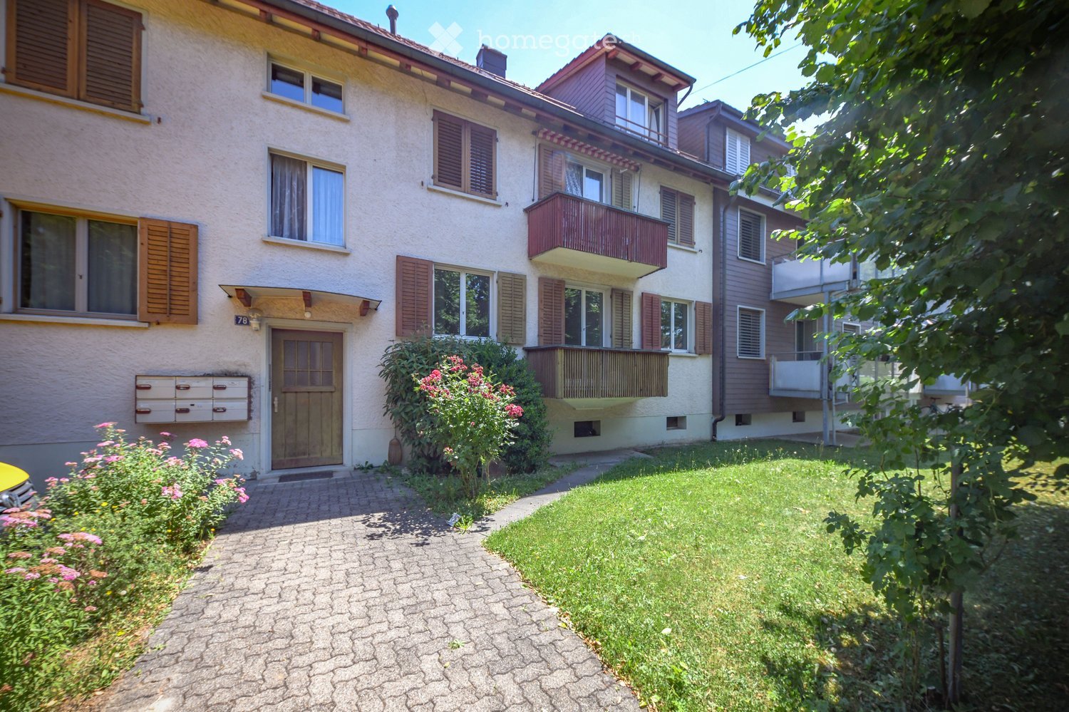 Two-story residential building with beige facade, wooden shutters, and balconies. The entrance features a wooden door and a small garden with flowers and shrubs.