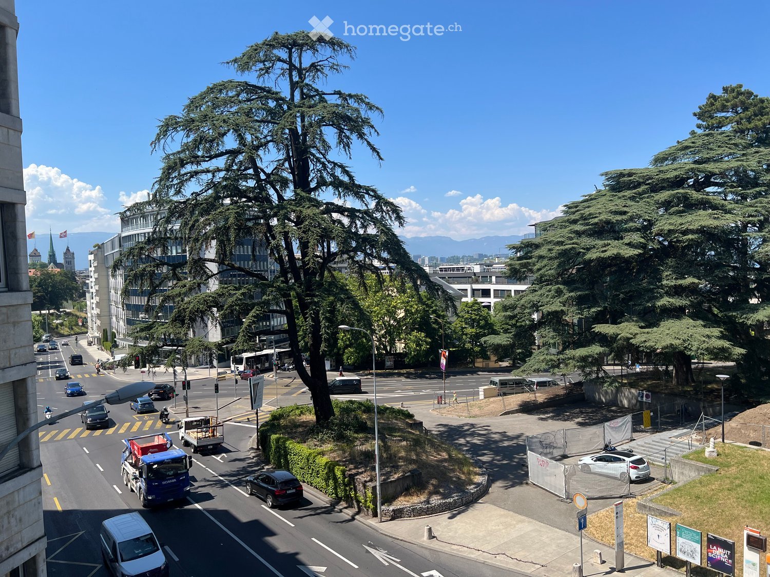 Urban street scene with multiple lanes of traffic, pedestrians, and surrounding buildings. Large trees and greenery are visible, with a clear blue sky and distant mountains in the background.