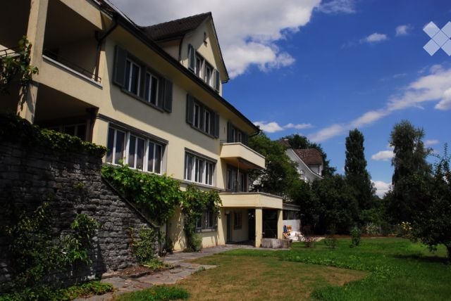 A large, multi-story residential building with cream-colored walls, multiple windows, and a stone staircase leading to the entrance. The building is surrounded by a well-maintained lawn and trees.
