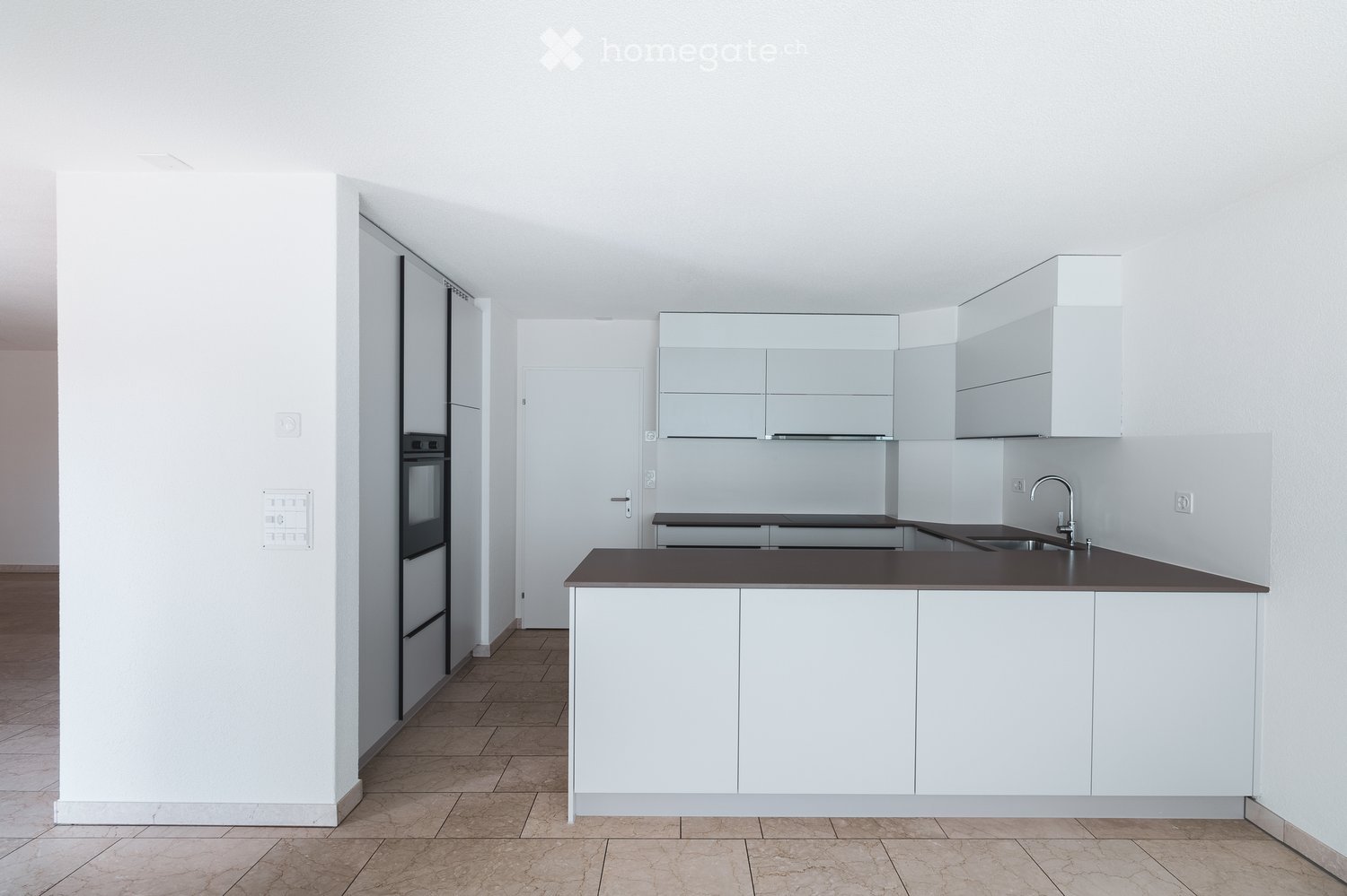 Modern, minimalist kitchen with white cabinets, dark countertops, a built-in refrigerator, and an oven. The space features a central island with a sink and faucet, and the floor is tiled.