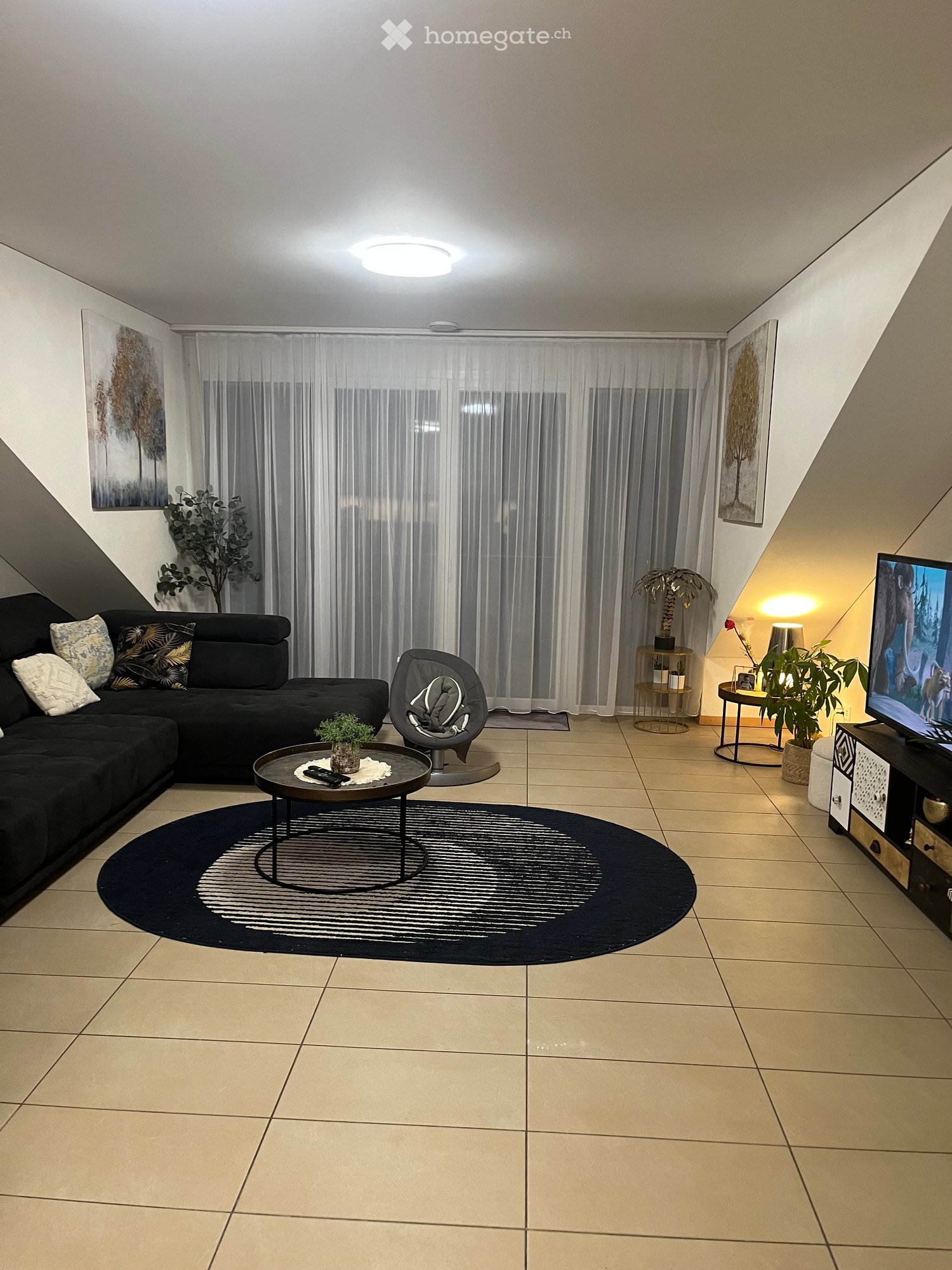 Modern living room with a black sectional sofa, coffee table, round rug, TV, and decorative plants.